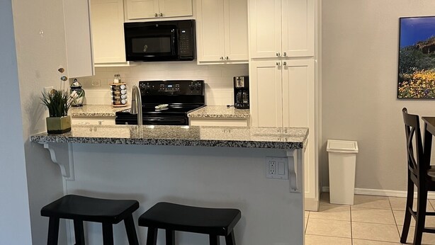 A kitchen with white cabinets, a black microwave, a black stove, and a granite countertop. There are two black stools and a trash can. - Gated Community, Poolside, Newly Remodeled 2bed Condo (Phoenix)