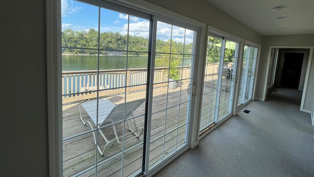 A room with large sliding glass doors leading to a wooden deck with a view of a lake and trees.