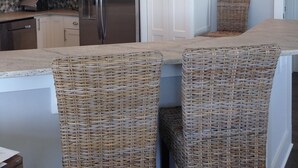 A modern kitchen with white cabinets, a stainless steel refrigerator, wicker chairs, and pendant lighting. - Newer Ocean View home w/Pool.....Walk to Ocean and Intercoastal (Oak Island)