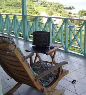 Un fauteuil à bascule en bois avec un coussin à motifs floraux, une petite table en bois sur laquelle se trouve un ordinateur portable, et en arrière-plan, une vue sur la verdure et une plage.