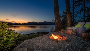 Ein Sitzbereich am Seeufer mit Feuerstelle, Holzbänken und Blick auf den See und die Berge im Abenddämmerung.