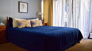 Cama bem arrumada com roupa de cama azul e almofadas estampadas, mesa de cabeceira com abajur e quadro pendurado na parede.