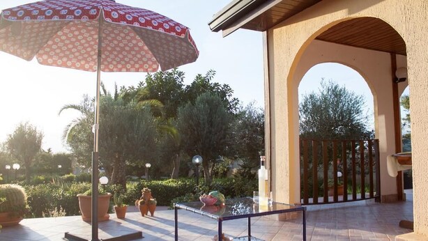 Ein Terrassenbereich im Freien mit einem Glastisch, einem gemusterten Sonnenschirm, Blumentöpfen und einem hölzernen Bogen.