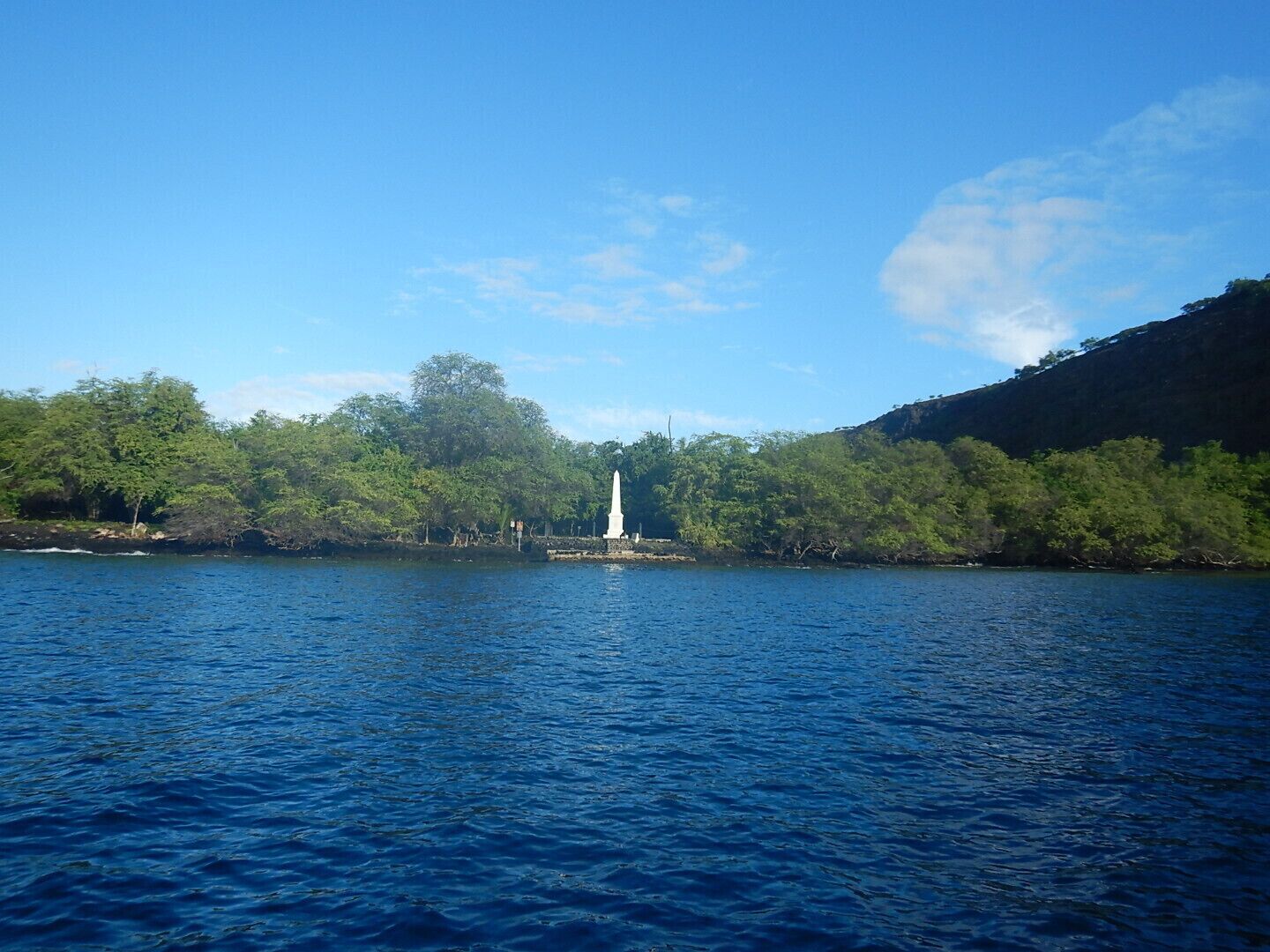 A calm lake with a white monument in the center, surrounded by lush greenery and a clear blue sky.