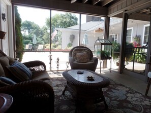 Un patio soleado con muebles de mimbre, una puerta de cristal que da al exterior y vistas a una casa y jardín.
