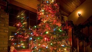Ett mysigt rum med ett stort julgran som är dekorerat med ljus och julpynt, omgivet av inslagna presenter och en jultomtefigur.