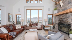 A spacious living room with leather sofas, a fireplace, large windows with mountain views, and a stone accent wall. - Big Sky Vacation Rentals: MMH 5 Derringer (Big Sky)
