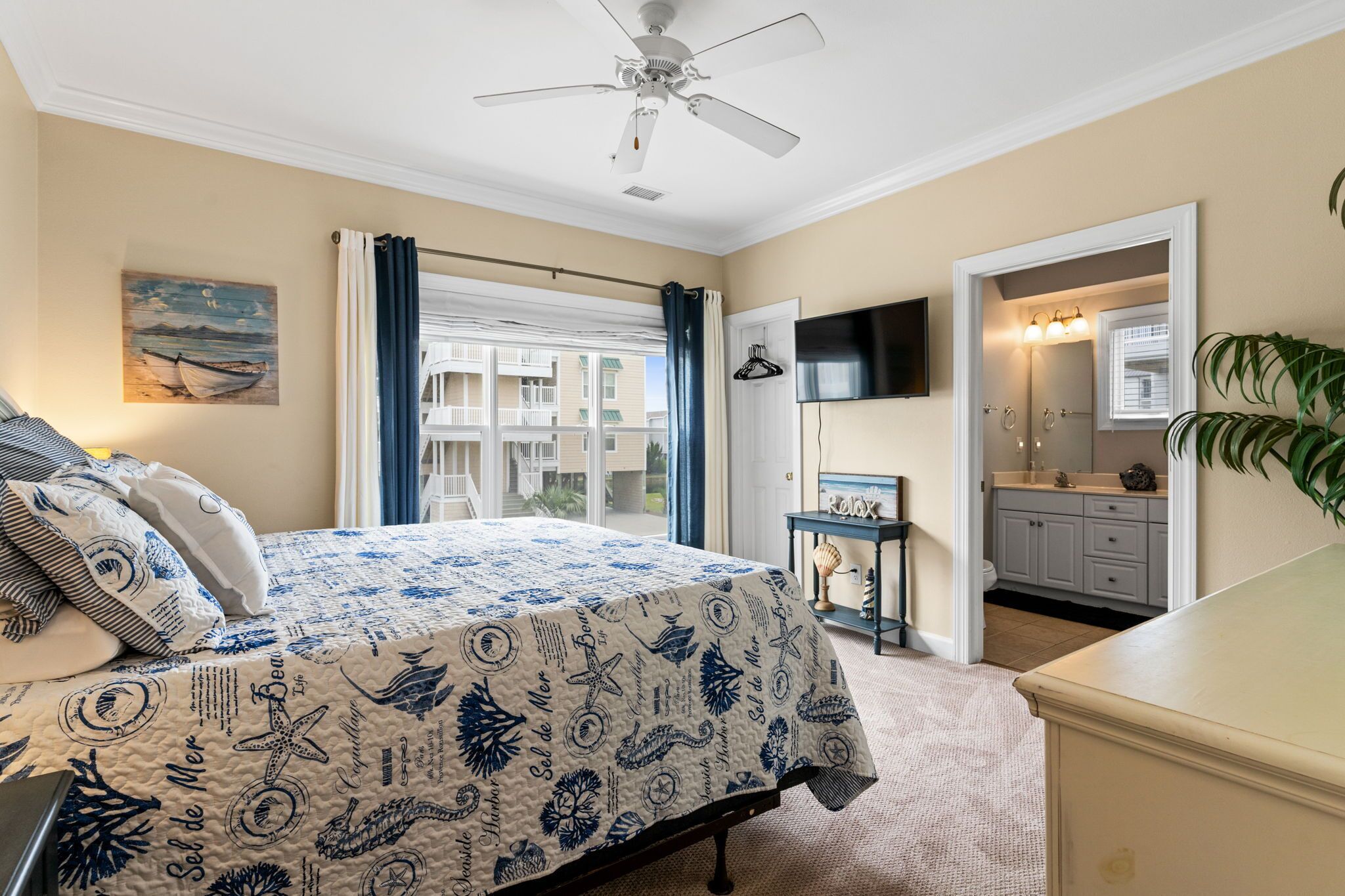 A bedroom with a bed, a ceiling fan, a painting, a window with curtains, a view of a balcony, and a bathroom with a mirror and sink.