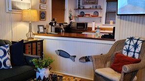 A cozy living room with a wooden coffee table, a wicker armchair, and a sofa. A kitchen area is visible through an open doorway, featuring a microwave, stove, and a 'Welcome' sign. - HIDEAWAY at HAWKINS!East TX LAKEFRONT Cabin in the Trees-Firepit-Private dock🐟 (Hawkins)