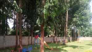 A playground with a slide, swings, and a climbing structure surrounded by trees and a brick wall. - Forest Breeze Gardens (Nairobi)