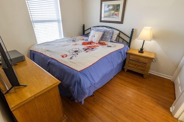 Un dormitorio con cama, cómoda de madera, mesita de noche con lámpara y un cuadro enmarcado en la pared.