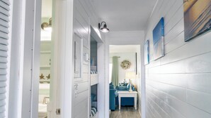 Un pasillo con paredes blancas, una luz en el techo y una puerta que da a una habitación con un sofá azul y una mesa blanca.