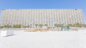 Un hotel grande con varios pisos y balcones, una playa de arena al frente y palmeras.