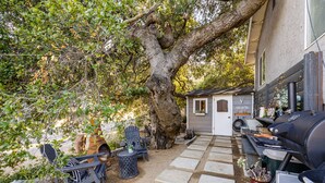 A backyard with a large tree, a barbecue grill, a small shed, and outdoor seating. - The B’s Hive Cottage (Newbury Park)