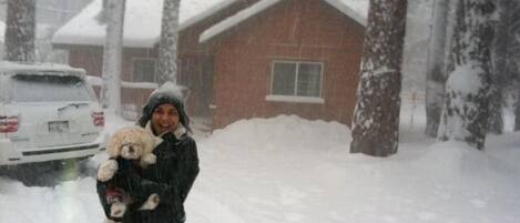 Una persona sosteniendo un perro en un entorno nevado al aire libre, con una casa y un automóvil al fondo.