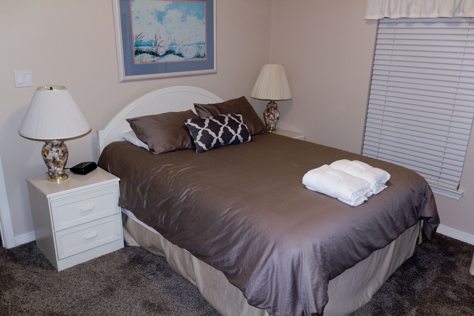 A neatly made bed with a brown bedspread, two pillows, and a folded white towel. There is a nightstand with a lamp and a telephone. A painting hangs on the wall above the bed.