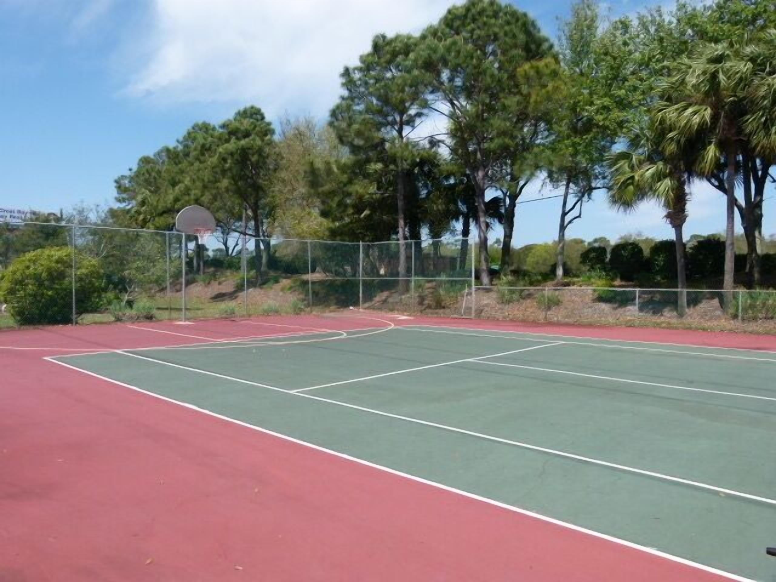 An outdoor tennis court with a basketball hoop, surrounded by trees and a fence.