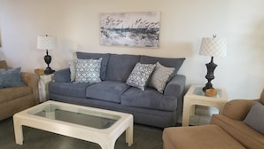 Una sala de estar con un sofá gris, cojines estampados, una mesa de centro de vidrio y dos lámparas sobre mesitas.