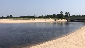A riverbank with a sandy beach, a bridge, and trees in the background. - Quaint beautiful cabin on the southwest shore on AuTrain Lake (AuTrain)