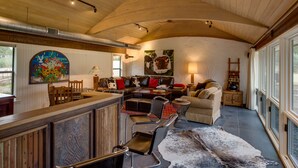 A spacious living area with a high ceiling, a wooden bar, leather furniture, and large windows. - Rio de Arcadia - Two Riverside Homes, Pool & Hot Tub (Kerrville)