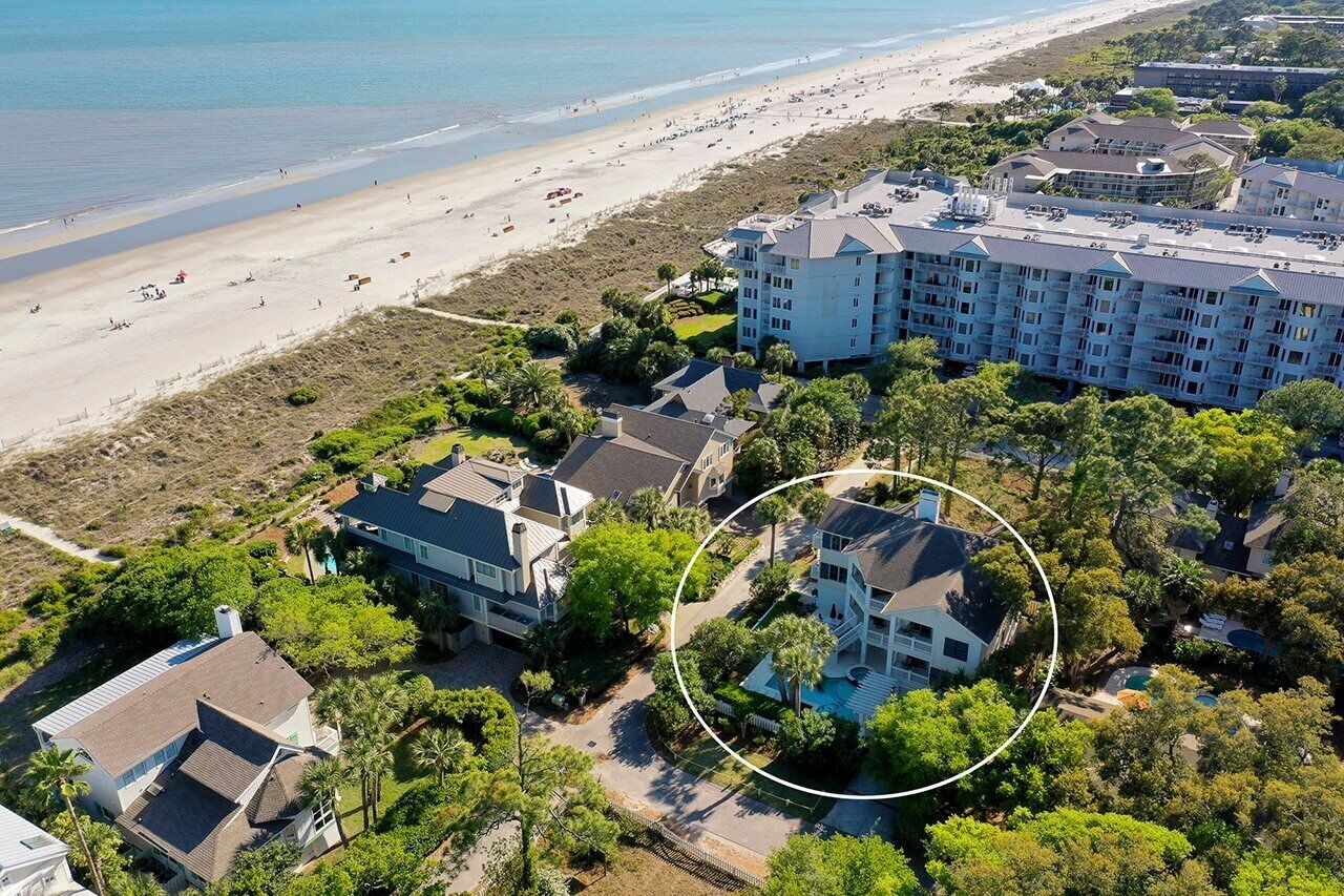 A coastal hotel with multiple buildings, a beach in the background, and a pool surrounded by lush greenery.