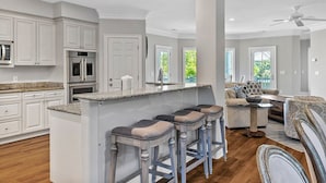 A spacious kitchen with white cabinetry, a central island with stools, a dining area with a sofa and table, and a ceiling fan. - Stunning Ocean View Vacation House with Private Pool, 1 Min From Beach! (Hilton Head Island)