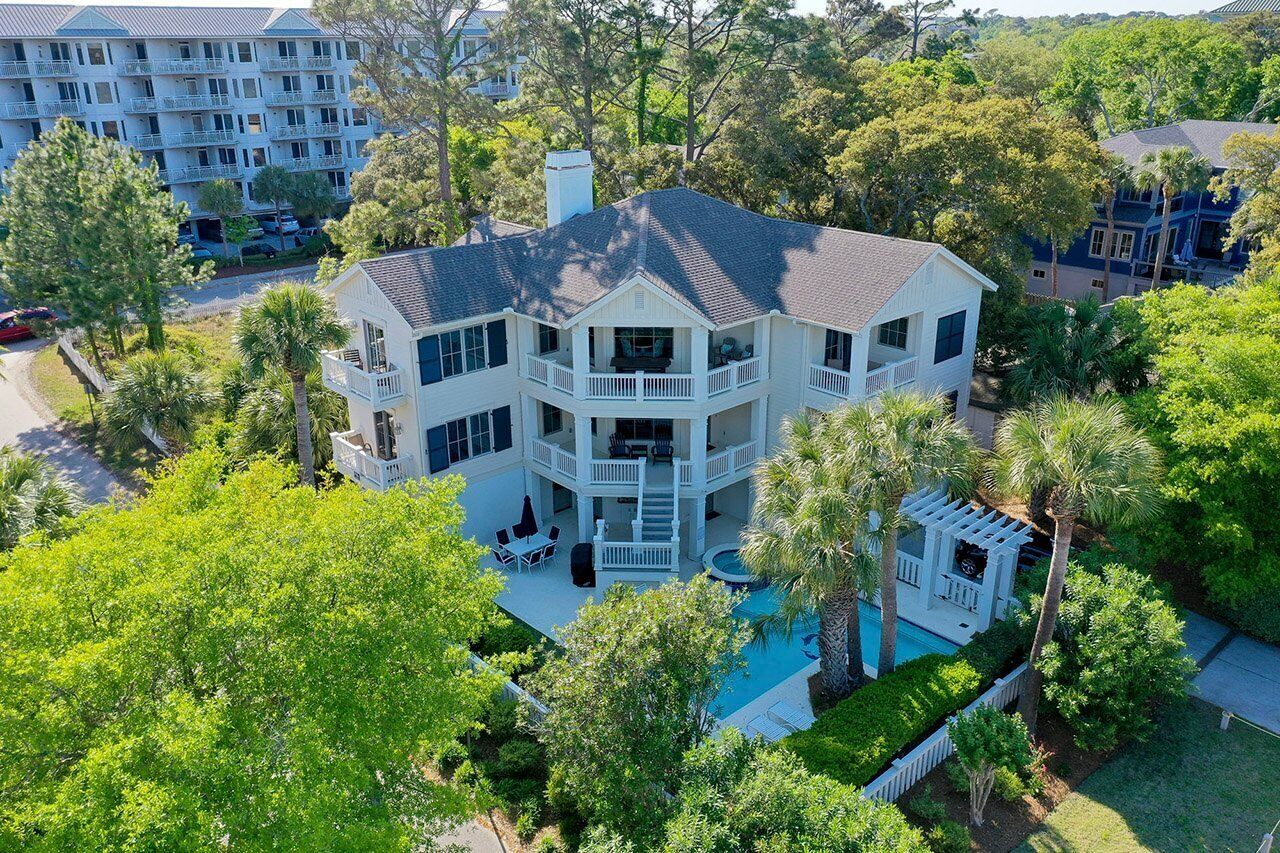 A large, multi-story house with a pool, surrounded by lush greenery and other buildings.