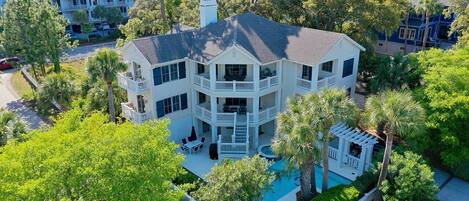 A large, multi-story house with a pool, surrounded by lush greenery and other buildings.