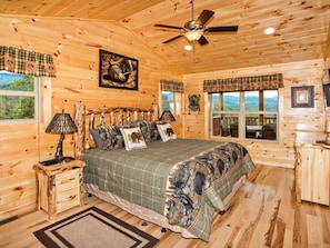 A log cabin bedroom with a bed, a ceiling fan, a window with a view, and a wooden nightstand.