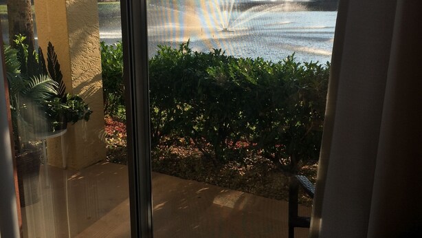A view through a glass door to a balcony with a water fountain, greenery, and buildings in the background.