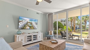 A living room with a flat-screen TV, a white entertainment unit, a beige sofa, a wooden coffee table, and a large sliding glass door leading to an outdoor patio with furniture. - Waterscape C200 ~ Ground Floor 3BR ~ Easy Access to Waterpark (Fort Walton Beach)