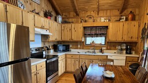 Une cuisine rustique aménagée dans une cabane en bois, avec un réfrigérateur, un four à micro-ondes, une cuisinière et un four, tous en acier inoxydable. On y trouve une table à manger en bois avec des chaises, une fenêtre ornée d’un rideau à motifs d’ours, et divers ustensiles de cuisine rangés sur des étagères.
