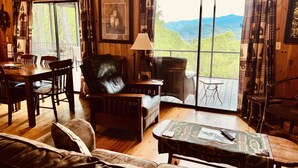 L’intérieur chaleureux d’un chalet avec des murs en bois, un coin repas, un fauteuil en cuir, un canapé et une grande fenêtre donnant sur une vue imprenable sur les montagnes.