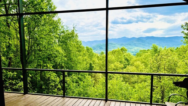 Une terrasse en bois avec une table en verre et un banc, offrant une vue sur un paysage montagneux boisé.