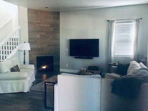 Una sala de estar con chimenea, un televisor montado en la pared, una escalera y una ventana con persianas.
