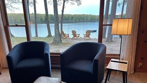 Ein Zimmer mit zwei dunklen Sesseln, einem kleinen Tisch und einer Lampe. Große Fenster bieten Blick auf einen See und Bäume.