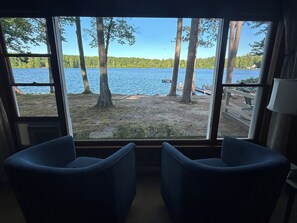 A room with a large window overlooking a lake, two chairs, and a lamp. - Leaning Pine: A Quiet Lakefront Cottage (Interlochen)