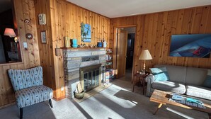 Ein gemütliches Wohnzimmer mit Kamin aus Stein, Holzwänden, einem gemusterten Sessel, einer Couch und einem Couchtisch.
