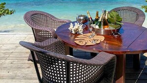 Eine Holzterrasse mit Korbstühlen und einem runden Esstisch, mit Blick auf das klare, blaue Meer und ein Boot in der Ferne.