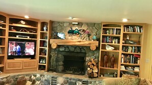 Una sala de estar acogedora con chimenea de piedra, estanterías de madera llenas de libros y objetos decorativos, un televisor y un espacio de descanso cómodo.