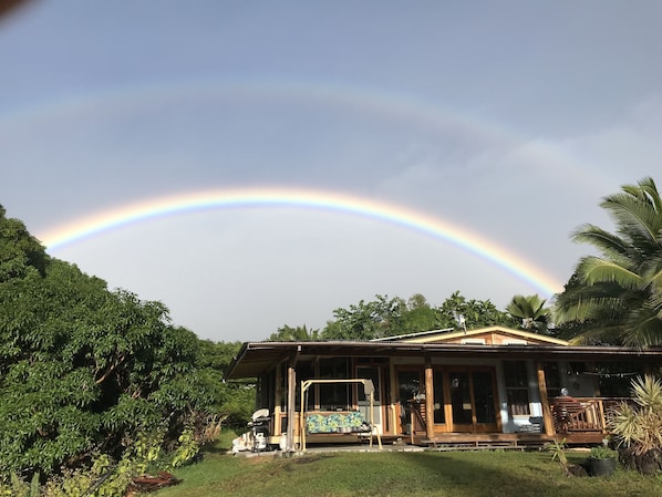 Ein Haus mit Veranda, umgeben von viel Grün und einem Regenbogen am Himmel.