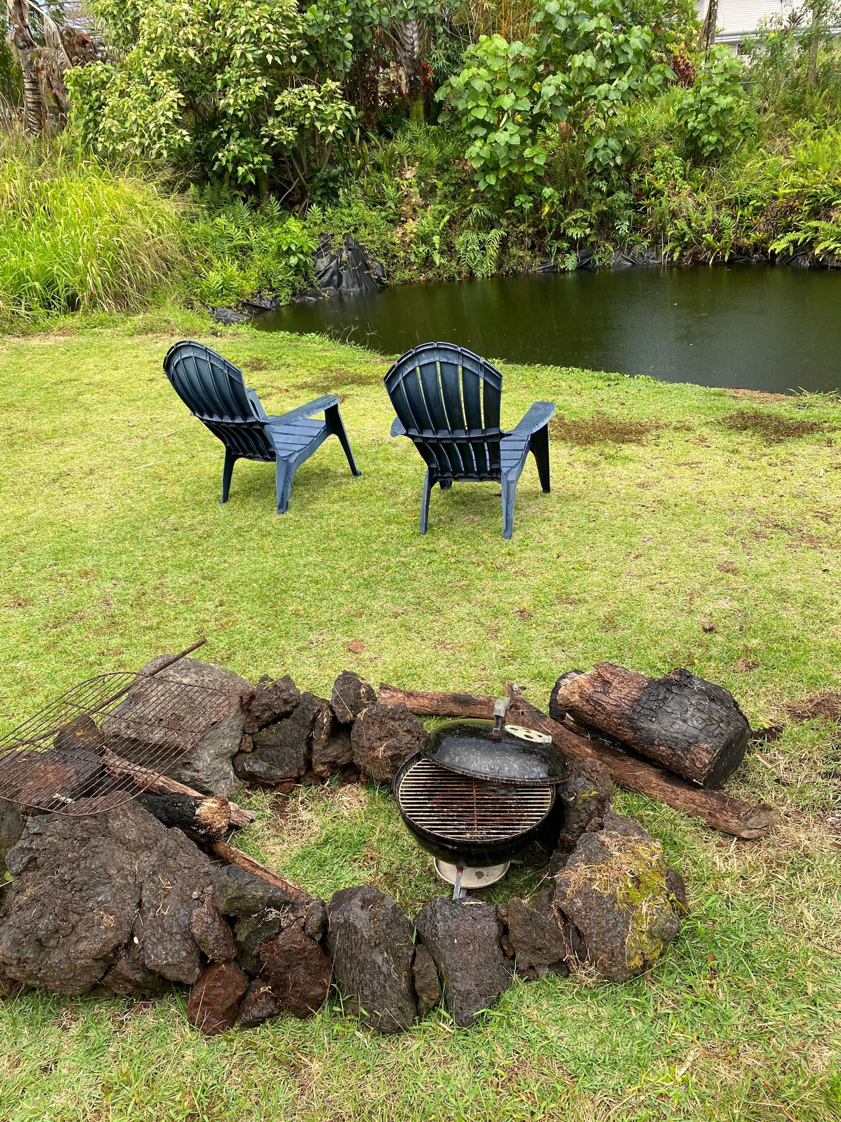 A grassy area with two Adirondack chairs and a stone fire pit with a grill.