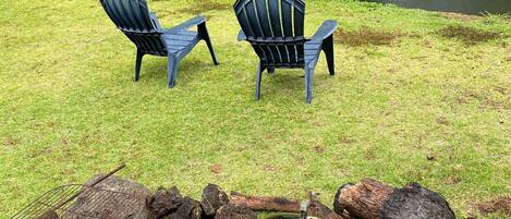 A grassy area with two Adirondack chairs and a stone fire pit with a grill.