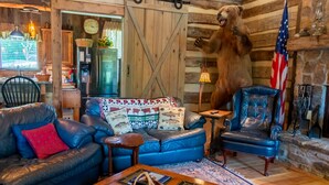 A rustic cabin interior with a leather sofa, a blue armchair, a coffee table with books, and a mounted bear head. - Vintage 1850's log house for rent with 185 acres  ,on a Creek ,  2miles trails . (Vanleer)