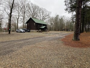 Un chalet rustique avec un toit végétalisé, entouré d’une allée en gravier et d’une zone boisée.