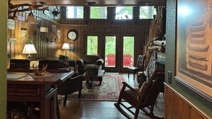 Interior rústico con paredes de madera, zona de comedor con mesa y sillas, chimenea y un ventanal con vistas a la vegetación.