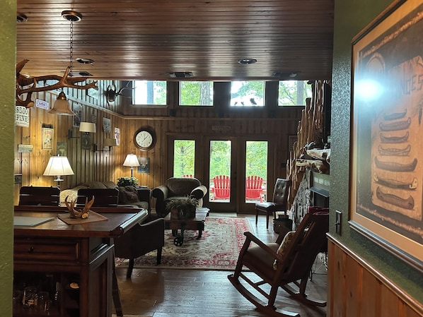 Interior rústico con paredes de madera, zona de comedor con mesa y sillas, chimenea y un ventanal con vistas a la vegetación.