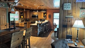 Un interior rústico con techos de madera, una zona de cocina, una barra con taburetes y una mesa de comedor con sillas.