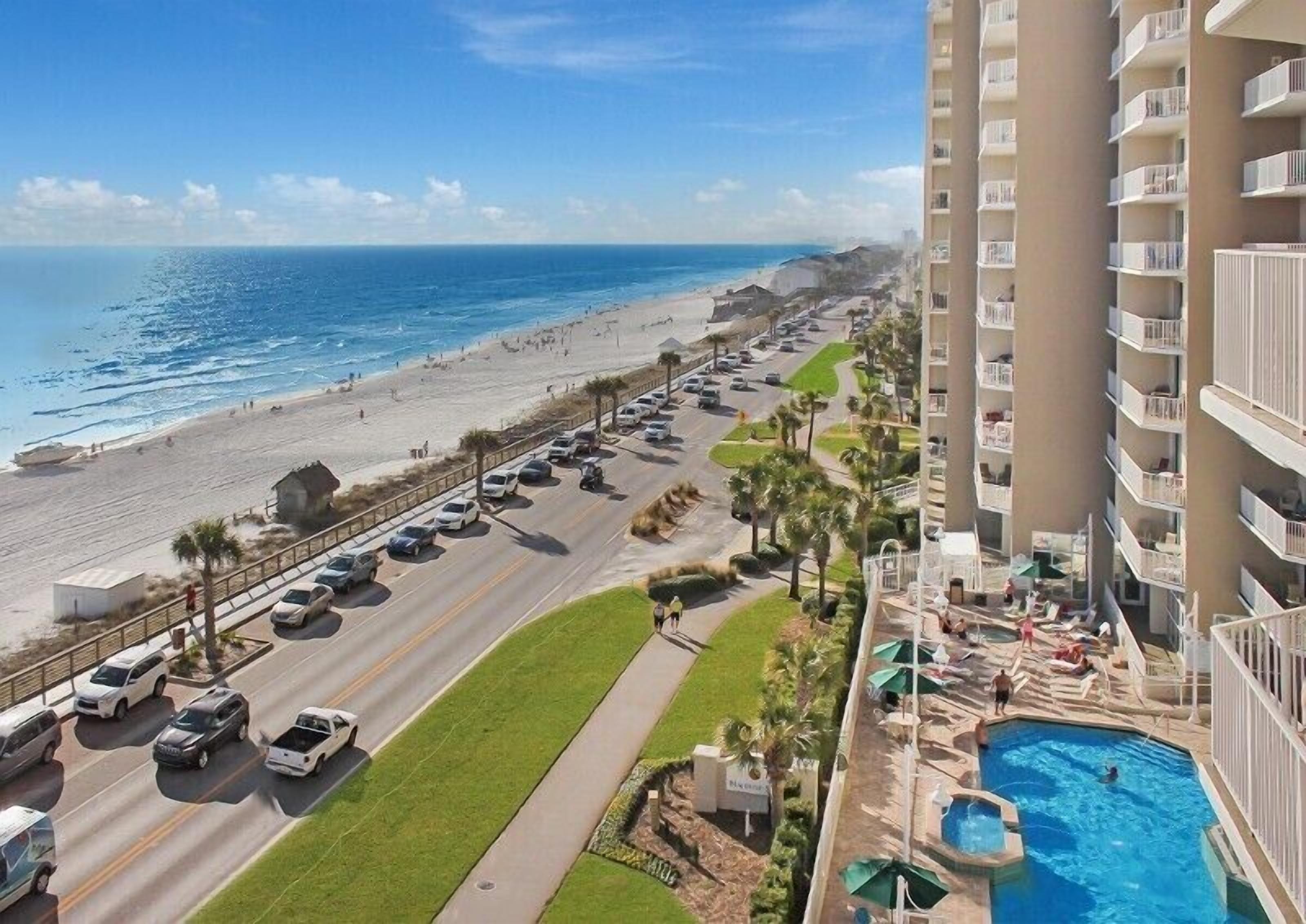 A hotel with a pool, a beach view, and a parking lot with cars.