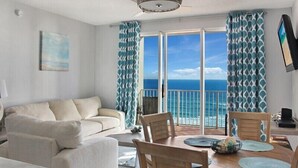 Een kamer met balkon met uitzicht op de oceaan, een bank, een eettafel met stoelen en een aan de muur bevestigde televisie.